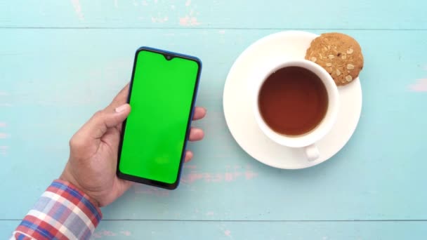 Junger Mann bedient Smartphone mit Tee und Keksen auf dem Tisch — Stockvideo