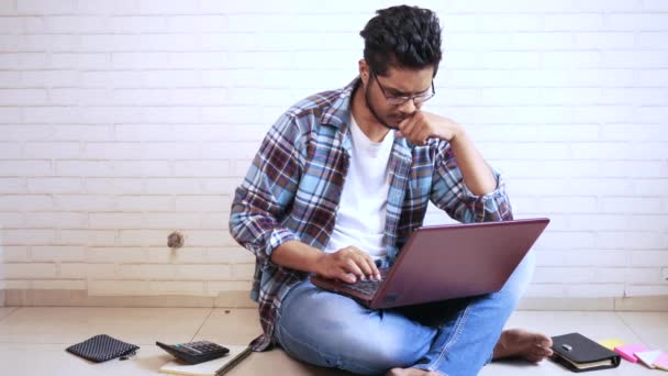 Серьезный молодой человек в повседневной одежде, сидящий на полу и работающий из дома — стоковое видео