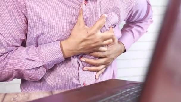 Jonge man lijden pijn in het hart en het houden van de borst, terwijl gezeten op het bureau — Stockvideo