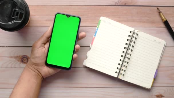 Молодой человек держит в руках смартфон с зеленым экраном на столе — стоковое видео
