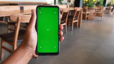 Birisi kafenin içinde yeşil ekranlı bir akıllı telefon gösteriyor..