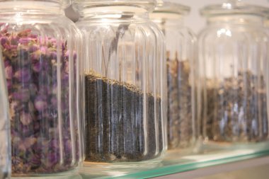 Several glass jars hold a variety of dried herbs and flowers on a shelf.