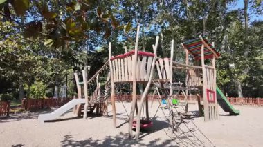 Çocuklar ağaçlarla çevrili ahşap bir oyun alanında oynuyorlar..