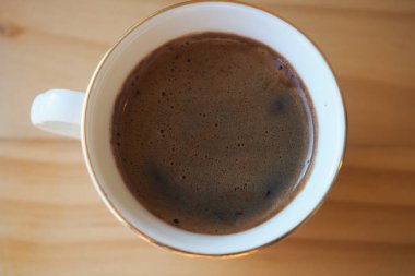 Koyu kahverengi kahve, pürüzsüz bir tahta yüzeyde duran beyaz bardağı doldurur..