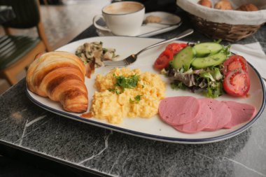Bir tabakta kruvasan, çırpılmış yumurta, salata ve kafede kahve bulunur..