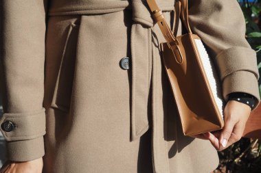 Bir kişi kahverengi bir ceket giyer ve dışarıda küçük bir çanta tutar..