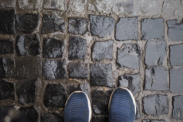 Yağmura batırılmış kaldırım taşları ayakkabının yanındaki suyun yansımasını gösteriyor..