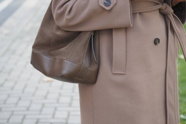 Bir kadın kaldırımda yürürken kahverengi bir çanta tutuyor..