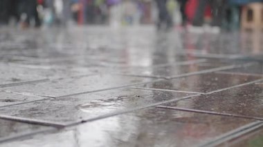 Kalabalık bir sokağa sağanak yağmur yağar, her yerde su birikintileri ve yansımalar yaratır..