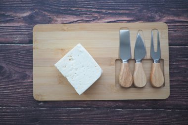 A block of cheese sits on a wooden board with three knives beside it.