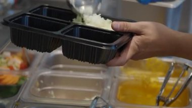 Bir insan, yoğun bir yerdeki kafeteryada yemek konteynırlarına pirinç servis eder..