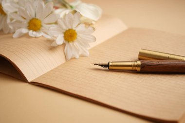 Yanında çiçekler olan açık bir defterin üzerinde bir dolmakalem duruyor..