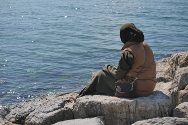 Bir kadın dalgaları izlerken su kenarında kayaların üzerinde dinleniyor..