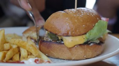 Bir insan patates kızartmasının yanında peynirli ve sebzeli bir burger keser..