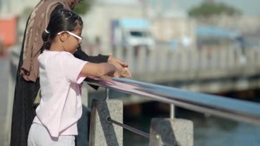 Bir kız suyun kenarında duruyor ve bir ebeveyn yakınlarındayken etrafa bakıyor..