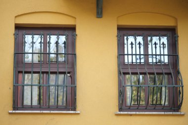 Güneş ışığında sarı bir duvarda demir parmaklıklı iki pencere görülüyor..