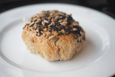 Yuvarlak bir ekmek rulosu beyaz bir tabakta ve yüzeyinde tohumlar var..