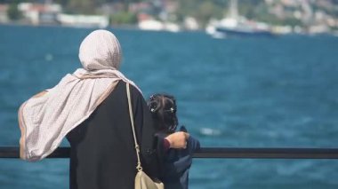 Bir anne ve çocuk korkulukların yanında durur ve suyu izlerler..