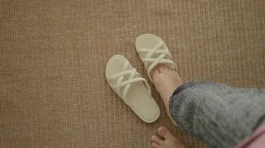 Kapalı alanda paspasın üzerinde terliklerin yanında yalınayak yürüyen bir insan..