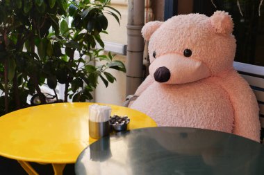 Büyük pembe bir ayı dışarıdaki yuvarlak bir masada tek başına oturur. Yakınlarda bir bitki var..