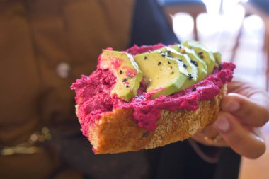 Bir insan pancar ezmesi ve avokado dilimleriyle kaplı bir dilim ekmek tutar..