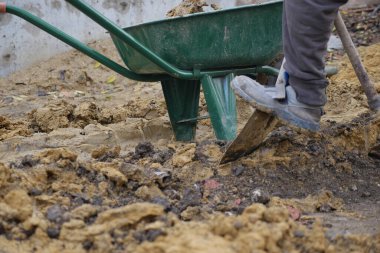 Worker uses shovel to move soil near a wheelbarrow in a construction area.
