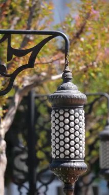 Bahçedeki metal bir kancaya asılı bir fener. Arka planda ağaçlar var.