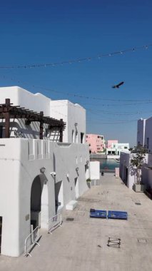 Katar Doha 2 Şubat 2025, mina bölgesindeki çöl kasabasında renkli mimari. İnsanlar, beyaz binalar ve deniz kenarında güneşli bir cadde boyunca yürüyorlar..