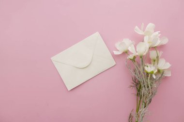 Pembe arka planda beyaz çiçeklerin yanında bir zarf duruyor..