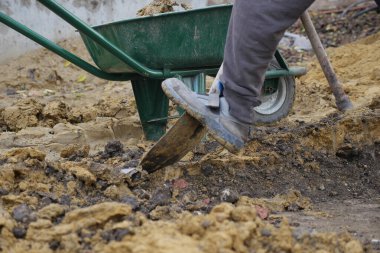 Bir işçi kazmak için el arabası kullanırken toprakta durur..