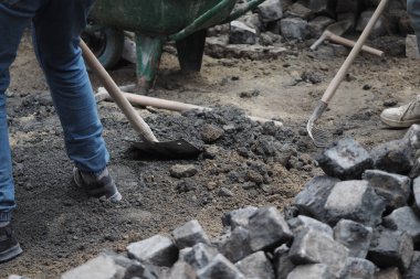 İki kişi inşaat alanında kürek ve tırmıkla kazı yapıyor..