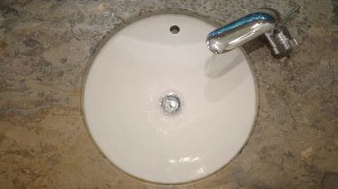 Musluktan yuvarlak bir lavaboya su akıyor. Lavabo beyaz malzemeden yapılmıştır..