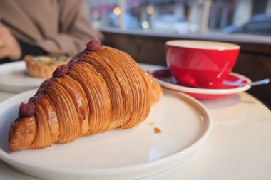 Bir kruvasan kahve dolu kırmızı bir bardağın yanında bir tabakta oturur..