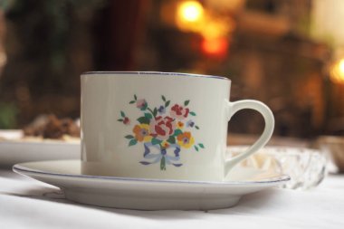 Arka planda çiçekler ve yiyecek olan beyaz bir masa örtüsünün üzerinde bir fincan duruyor..