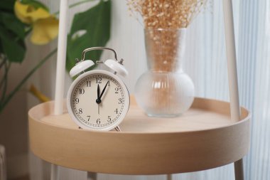 Bir saat bir bitki ve kuru çiçekli bir vazonun yanındaki masada oturur..