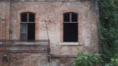 Eski bir tuğla bina boş pencereler gösteriyor ve etrafında bitkiler yetiştiriyor..