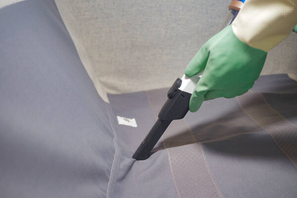 A worker uses a vacuum to clean a sofa in a room during the day.