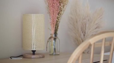 Masasında kurumuş bitkiler olan bir vazonun yanında bir lamba duruyor..