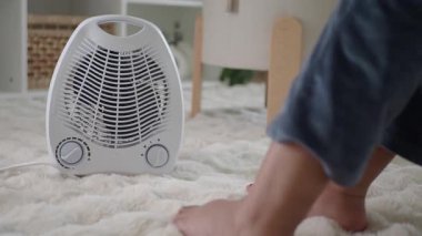 Bir insan otururken elektrikli ısıtıcıyla ayaklarını ısıtır..