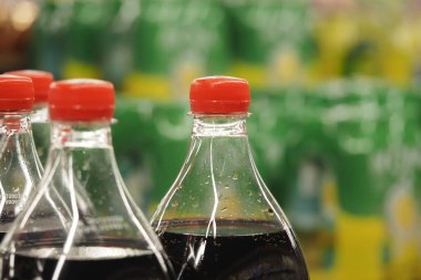 Soda şişeleri bir rafta, arkasında yeşil ve sarı şeyler var..