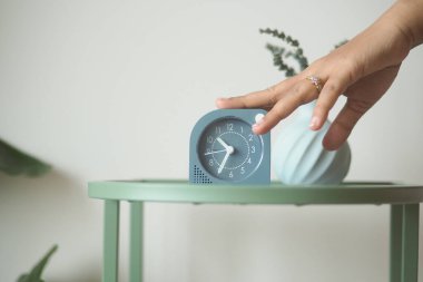 Bir el, vazonun yanındaki yeşil masadaki saati bitkilerle ayarlar..