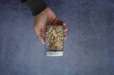 Bir el, fındık parçalarıyla dolu cam bir kavanozu gri arka planda tutar..