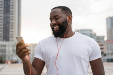 Genç yakışıklı adam cep telefonu kullanıyor, iletişim kuruyor, dijital ekrana bakıyor, sokakta yürüyor. Afro-Amerikalı adam akıllı telefon tutuyor, müzik dinliyor, açık havada gülümsüyor.