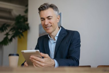 Modern ofiste online çalışan, dijital tablet okuyan yakışıklı bir iş adamı. Gülümseyen orta yaşlı adam online alışveriş yapıyor, evde oturuyor.  