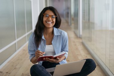 Afrika kökenli Amerikalı öğrenci, sınav hazırlığı, uzaktan öğrenme, eğitim konsepti. Güzel, gülümseyen bir kadın dizüstü bilgisayar kullanıyor, internette notlar alıyor, yerde oturuyor. 