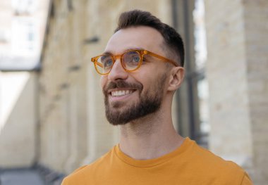 Yakışıklı, gülümseyen, sakallı, şık gözlüklü bir adamın yakın portresi sokağa bakıyor.