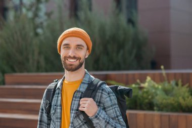 Sırt çantalı, sokakta yürüyen genç bir adamın portresi. Sıradan kıyafetler ve turuncu şapka giyen yakışıklı hippi dışarıda kameraya bakıyor.