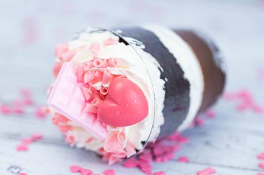 Kahverengi çikolatalı tatlı ve şeffaf bardakta beyaz krema. Pasta pembe çikolata ve kıvırcık bir kalple süslenmiş. Doğum günü pastası