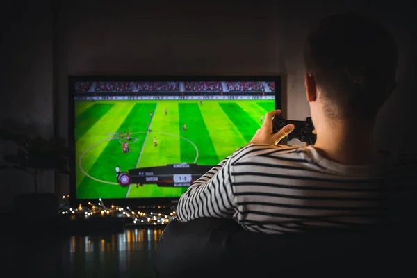 Young hipster man playing video game football soccer on console. gamer guy with gamepad controller holding Wireless joystick sitting on pouf at night. Back view. Fun entertainment at home isolation.