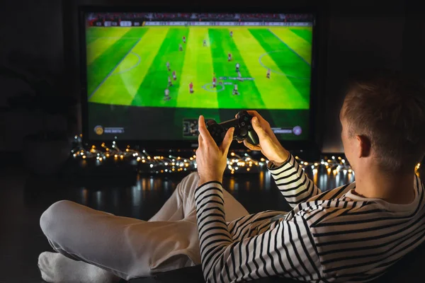 Young hipster man playing video game football soccer on console. gamer guy with gamepad controller holding Wireless joystick sitting on pouf at night. Back view. Fun entertainment at home isolation.